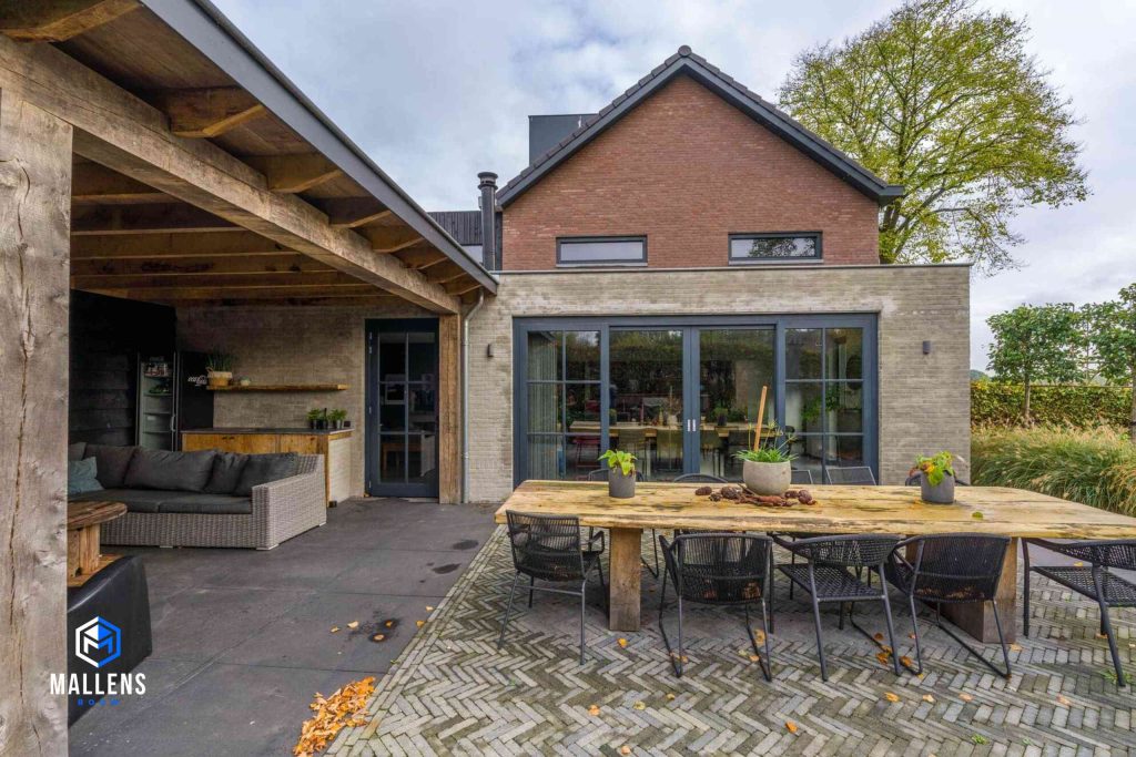 Je ziet de uitbouw met de grote zwarte serre deuren recht van voren. Voor de uitbouw staat een buitentafel met stoelen. Links zie je de overkapping met een loungebank.