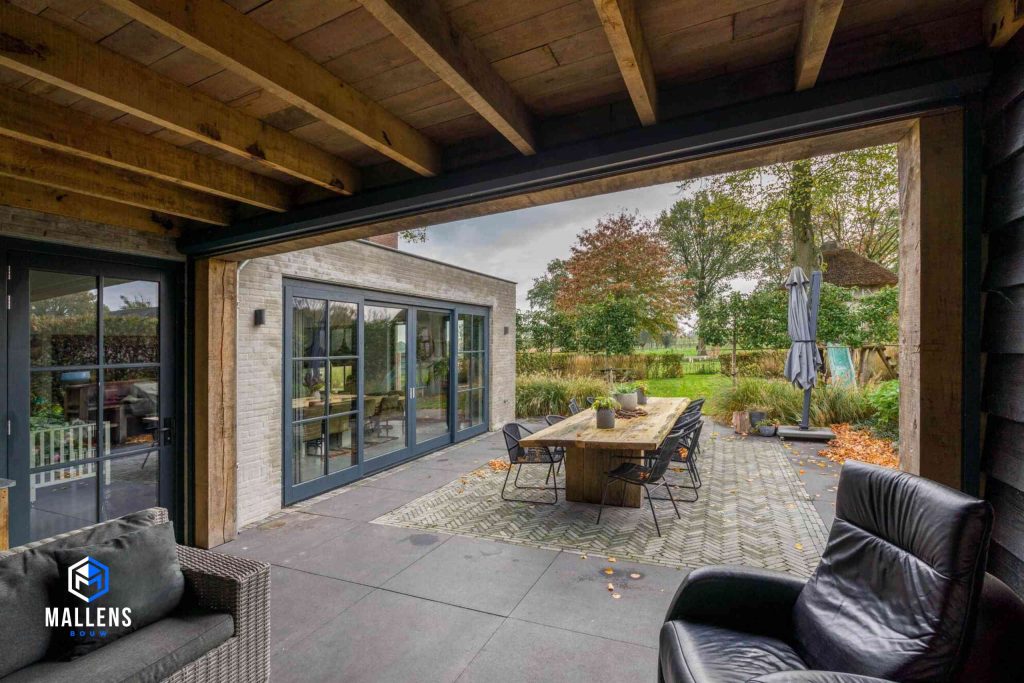 De overkapping met zicht op de achtertuin, waar een tafel met stoelen staan. Ook zie je de uitbouw met serre deuren.