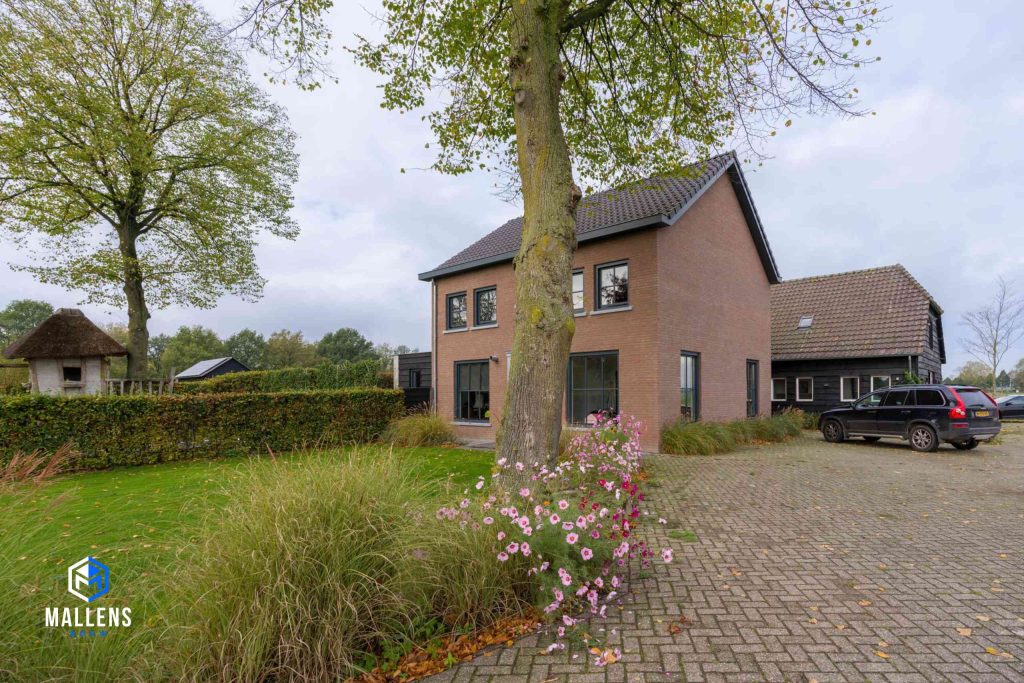 Eindresultaat van de renovatie van de vrijstaande woning. De woning van schuin voren gezien, met een grote boom voor het huis en roze bloemen.