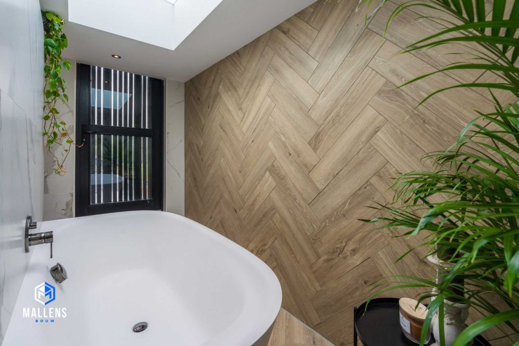 Een andere hoek van de badkamer met rechts een visgraat houten muur. De anderen muren zijn wit marmer. Er staat een zwart tafeltje met decoratie en je ziet een wit bad. In het plafond zit een raam.