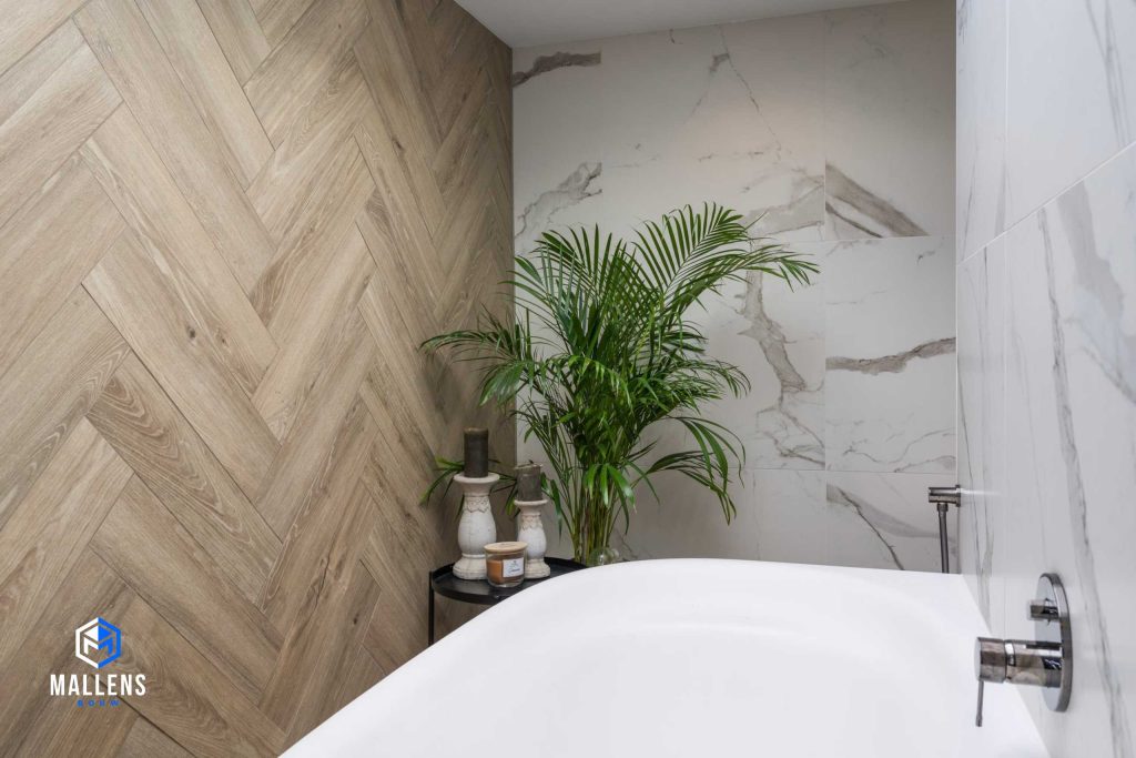 Een andere hoek van de badkamer met links een visgraat houten muur. De anderen muren zijn wit marmer. Er staat een zwart tafeltje met decoratie en je ziet een wit bad.