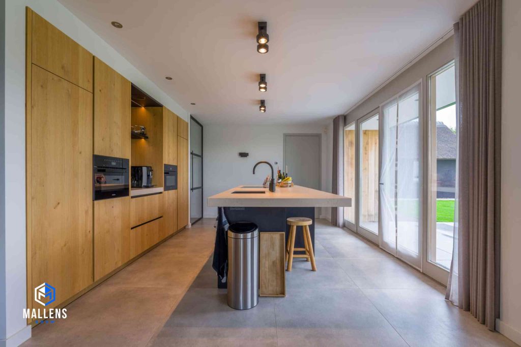De keuken met houten kasten en een keukenblok in het midden. Er komt veel licht naar binnen door de grote ramen en deuren.