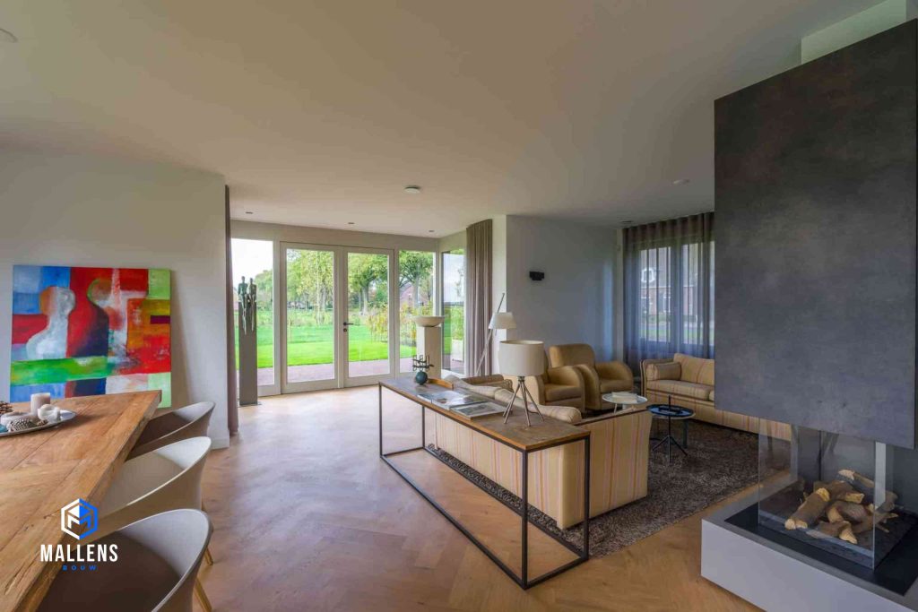 De zithoek met 2 gelige banken en 2 gelige loungestoelen. Er staat ook een poenhaard en je hebt zich op de tuin door middel van grote deuren en ramen.