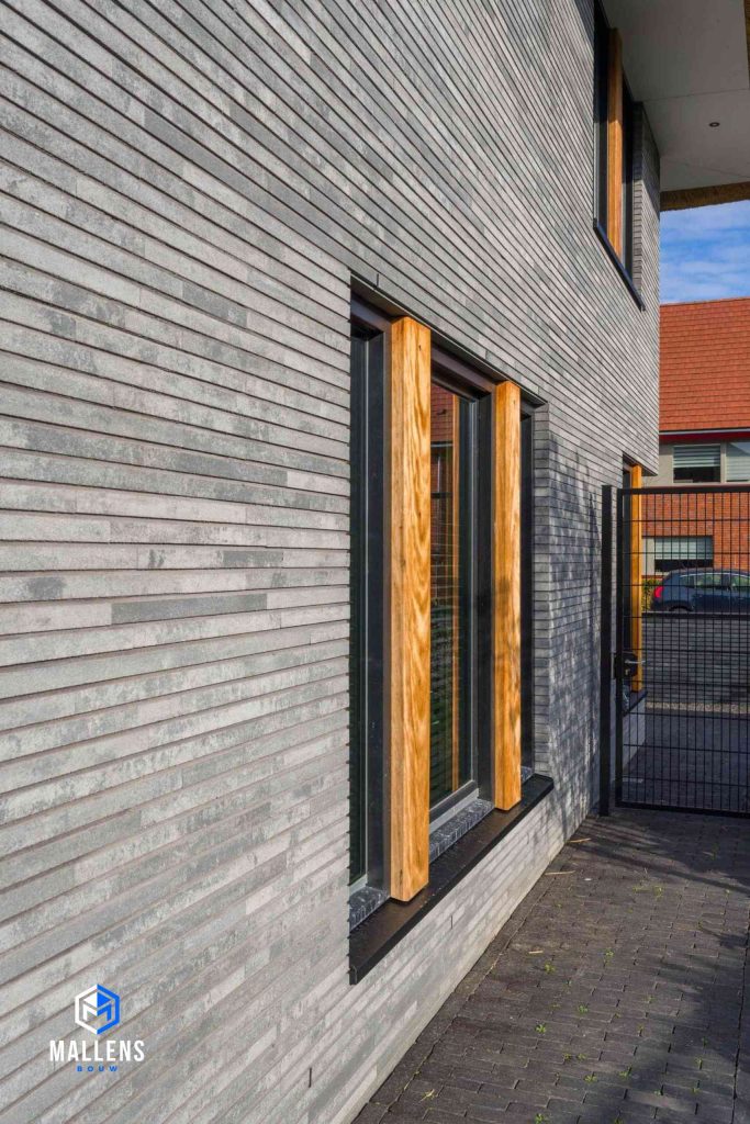 De grijze bakstenen muur met de zwarte kozijnen en houten balken bij het raam.