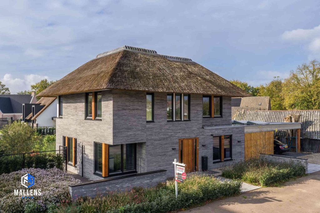 Het huis van schuin voren bekeken. De woning heeft veel grote ramen met zwarte kozijnen en houten accenten. Rechts aan de woning zit de garage.