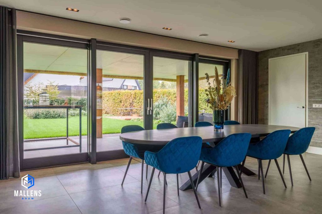 De eethoek van de woning met een grote tafel en 8 blauwe eetstoelen. Die staat voor een grote pui.
