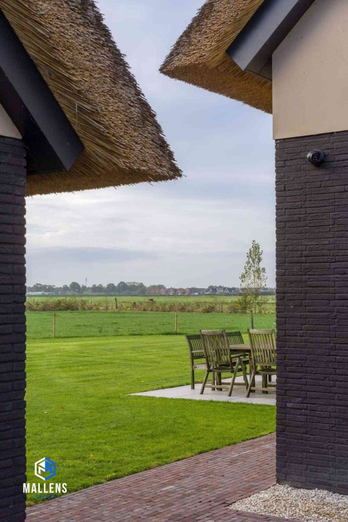 Je kijkt tussen de schuur en de woning door, waardoor je zicht hebt op de groene weilanden om het huis heen.