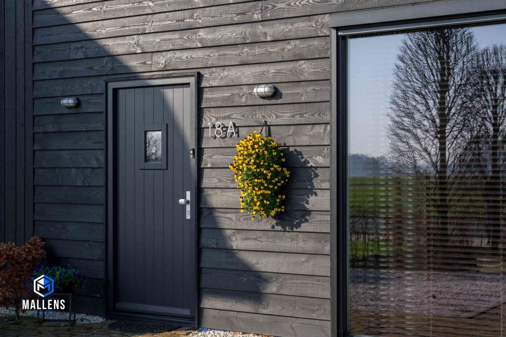 Detailfoto van de zwarte houten deur aan de voorzijde van de woning met daarnaast het huisnummer en een hangende bloempot met gele bloemen.