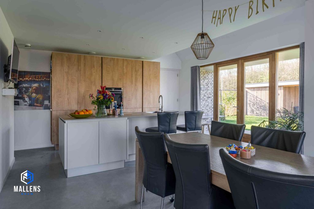 De keuken met houten kastjes, keukenblok met twee barstoelen en een eettafel met daaromheen zwarte leren stoelen.