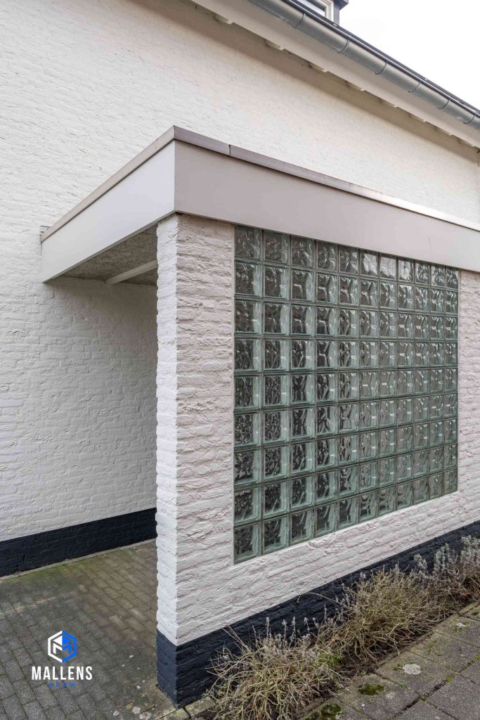 De muur van de overkapping die bij de deur van de schuur aanwezig is. In het muurtje zit een raampje met gehamerd glas.
