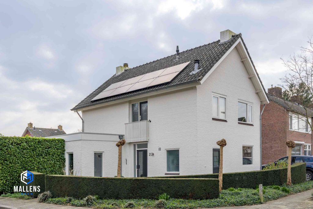 De woning in Oisterwijk gezien van schuin van voren. Het is een witte woning met zonnepanelen op het dak. Voor de woning zijn lage hegjes geplaatst en een drietal boompjes.