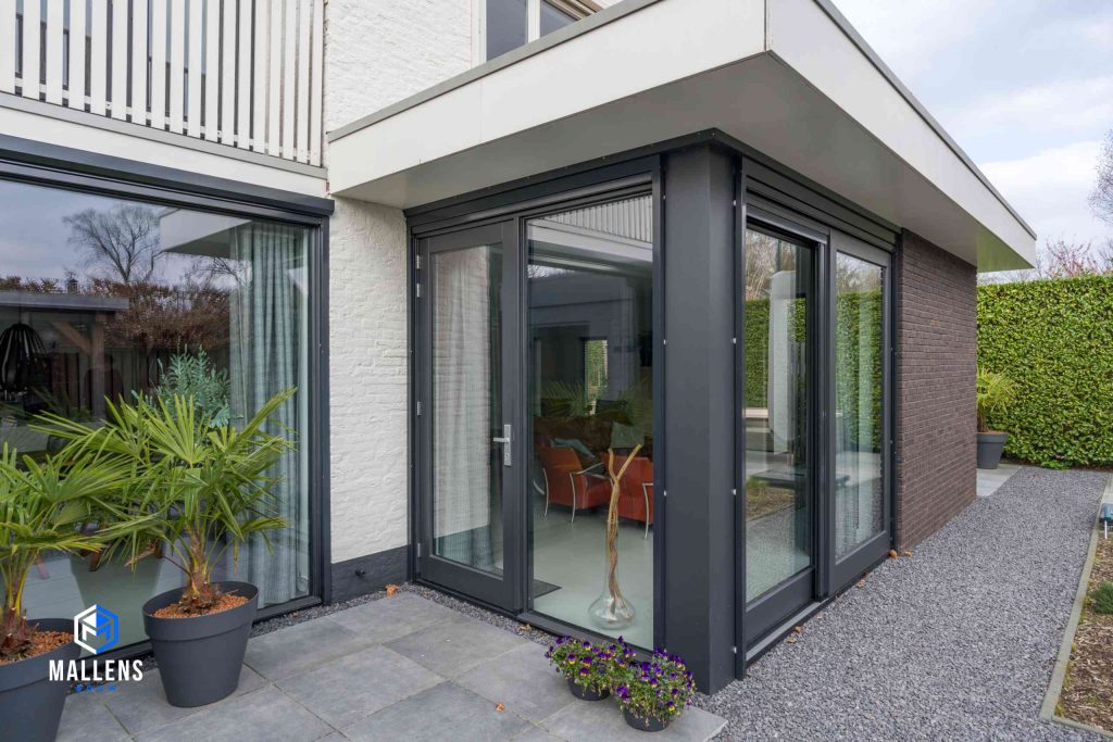 De uitbouw van de woning van dichtbij gezien, met grote ramen. De muren bestaan, in tegenstelling tot de woning, uit bruine bakstenen.