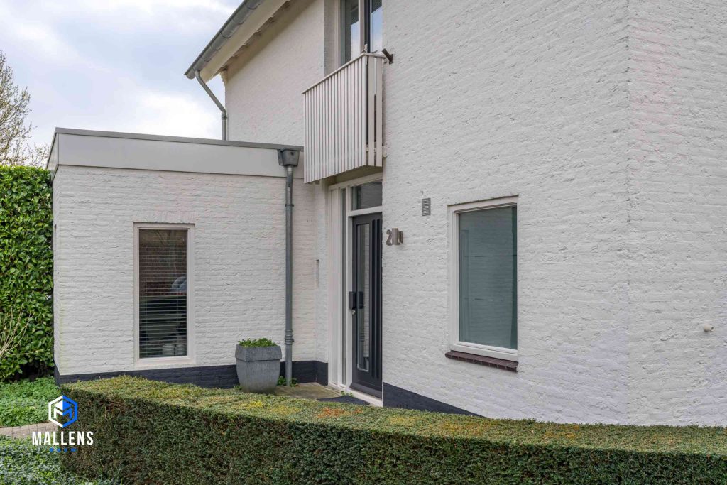 Je ziet de voorzijde van het huis met zicht op de voordeur. De woning heeft hier een witte uitbouw.