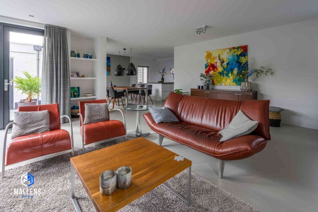 De zithoek van de woning met een rode leren bank en twee leren, rode loungestoelen. Er ligt een vloerkleed met daarboven een bijzettafeltje. Op de achtergrond hangt een kleurrijk schilderij.