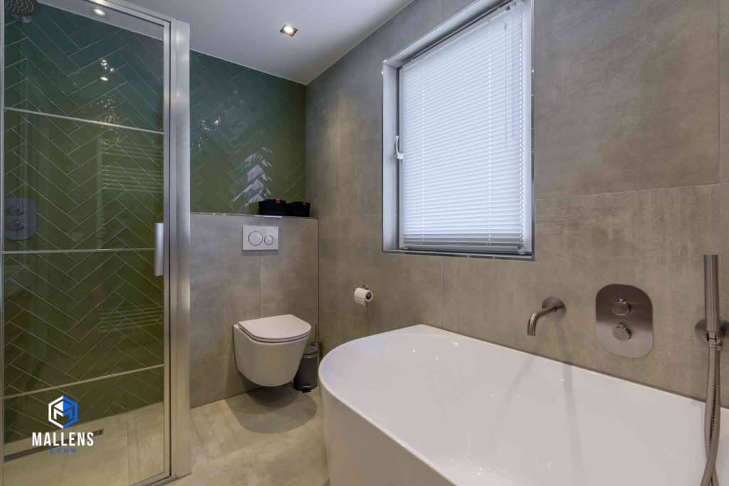 De badkamer met een zwevend toilet en ruim bad. Een gedeelte van de muur is betegeld met groene visgraat tegels. De rest met grijze vierkante tegels.