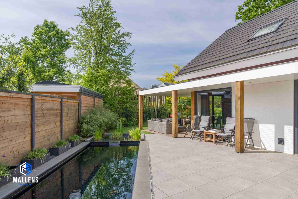 De tuin van de woning met een grote vijver en zicht op de veranda. De veranda wordt ondersteund door houten balken.