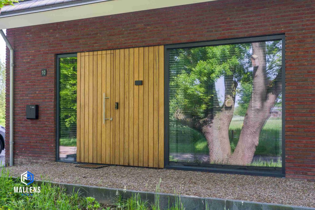 De houten voordeur van dichtbij bekeken. Het is een paneeldeur. Tegen de deur aan zit een groot raam met zwarte kozijnen.
