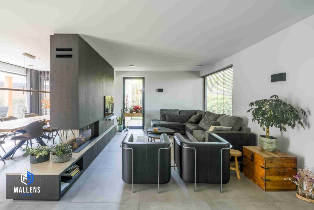 De woonkamer van de nieuwbouwwoning in Heukelom met een grote hoekbank en twee fauteuils. Een grote wand met kachel en televisie scheidt de woonkamer van de eethoek.