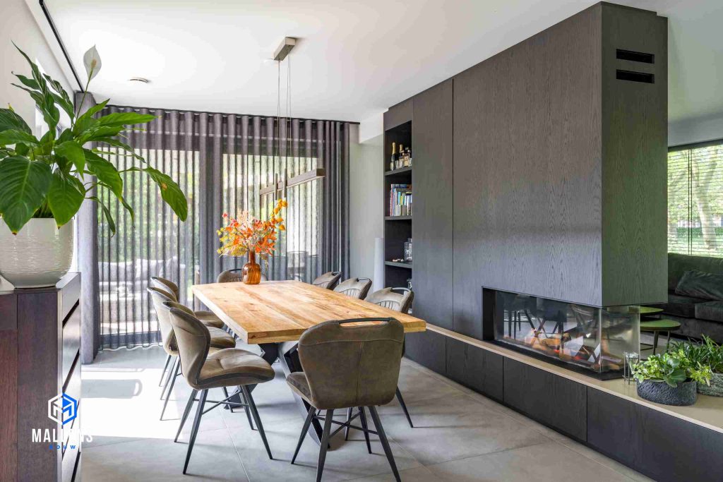 De eethoek met een houten tafel en 8 stoelen eromheen. Het staat voor een groot raam met doorschijnende gordijnen.